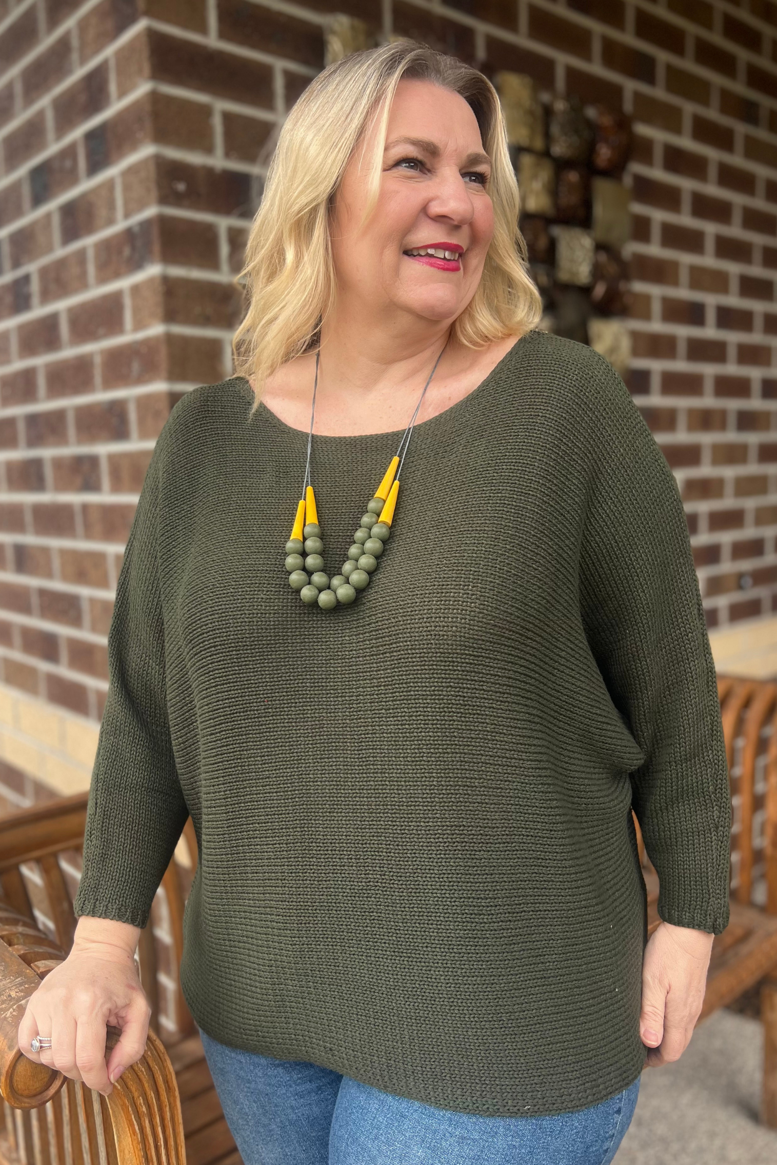 Dark green knitted boat necked top and necklace over jeans, standing on porch