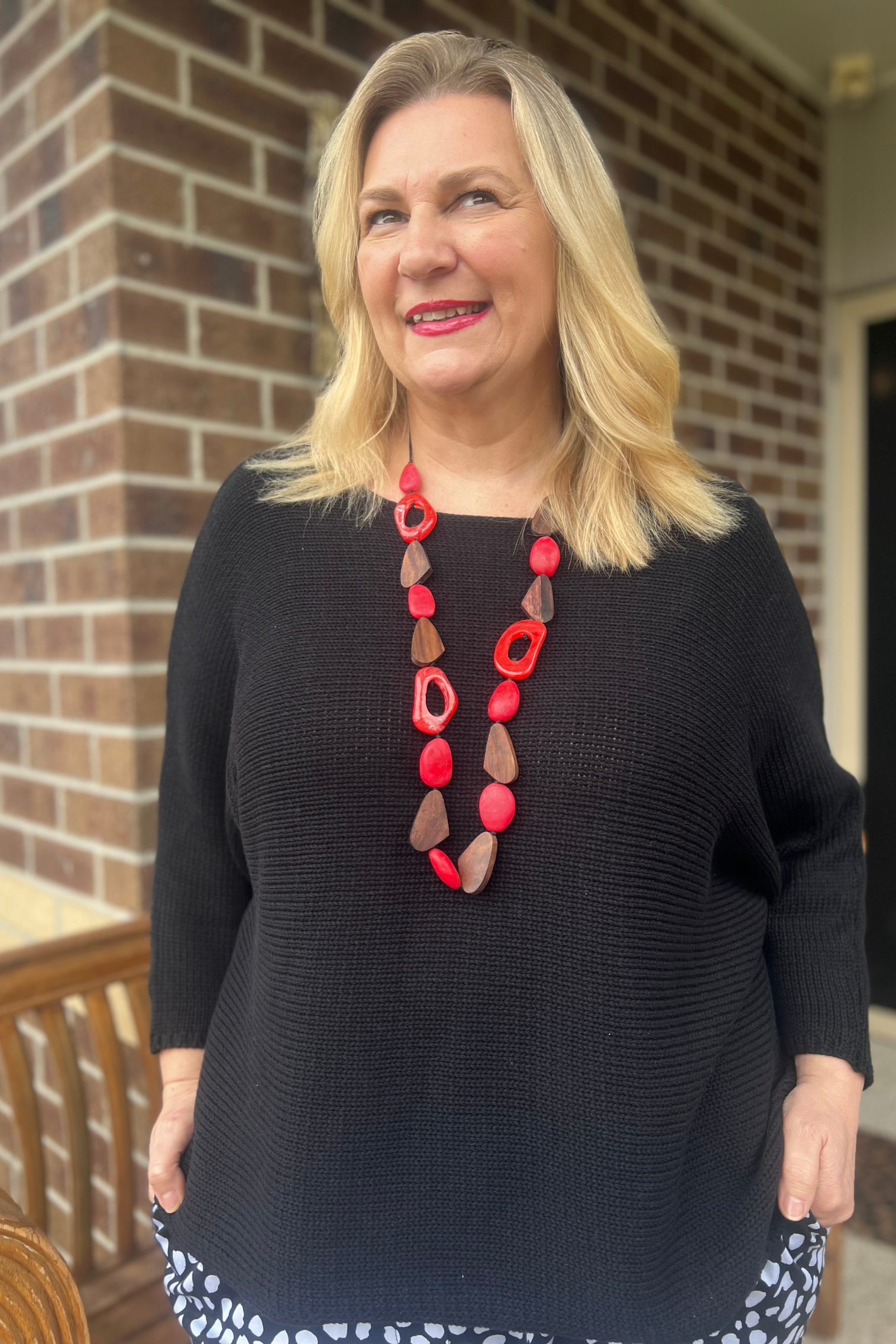 Kita Ku modelling a black boat neck sweater and red necklace on a porch setting. 