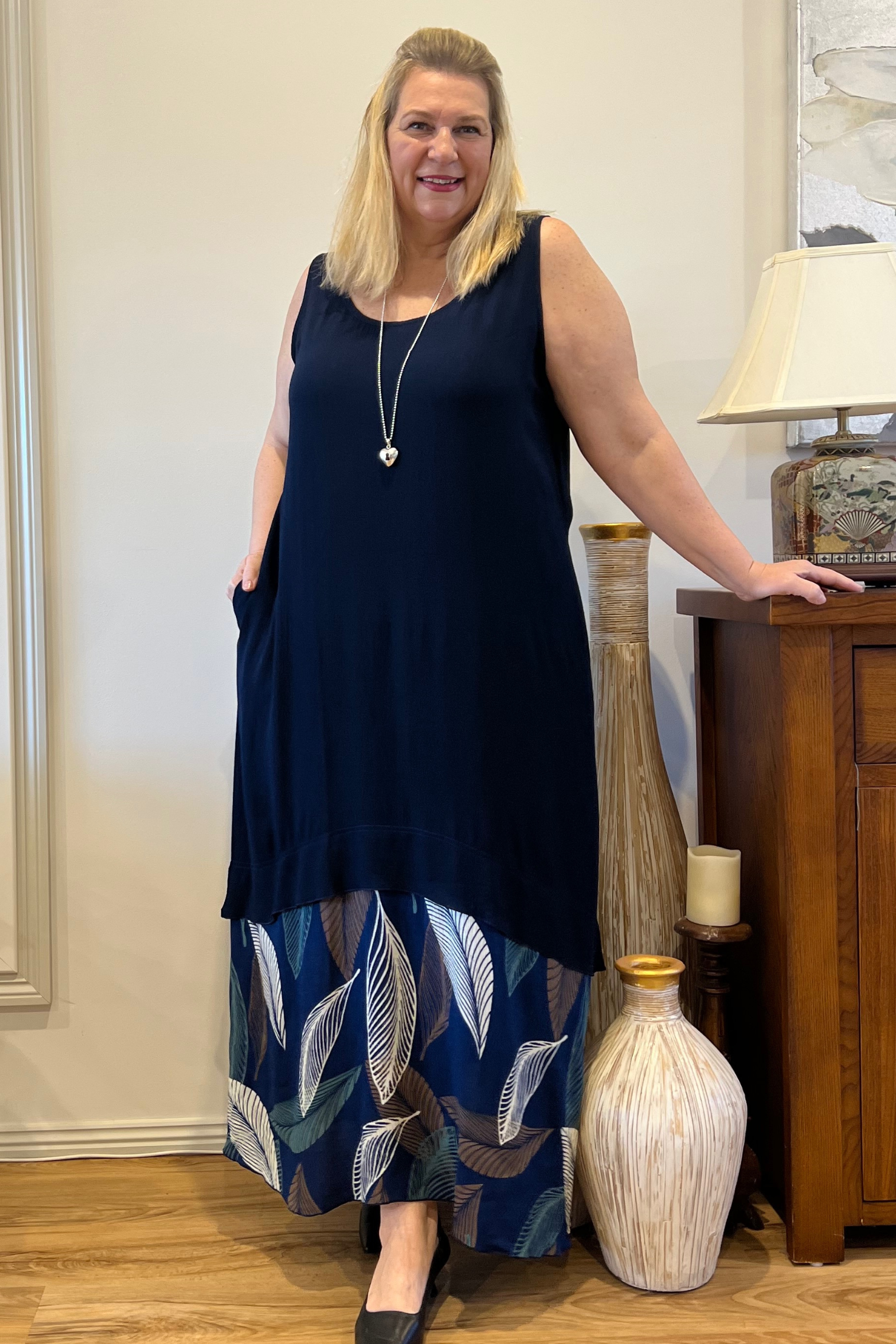 kita ku model wearing a maxi no sleeve navy dress with a combo panel on the bottom of assorted large leaves