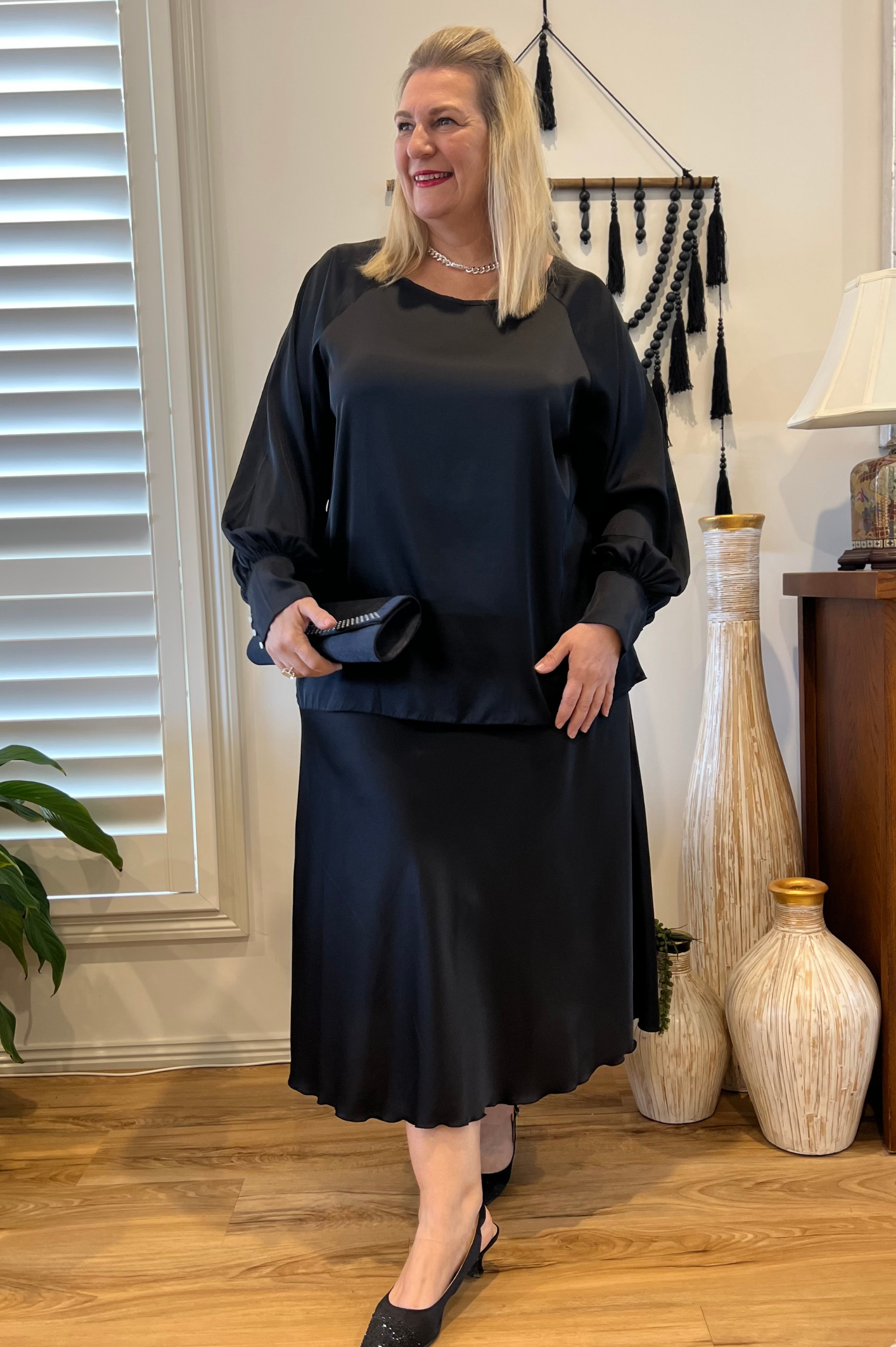 Kita Ku model wearing a black satin skirt and a matching long sleeve blouse with  a full sleeve on a long band.
