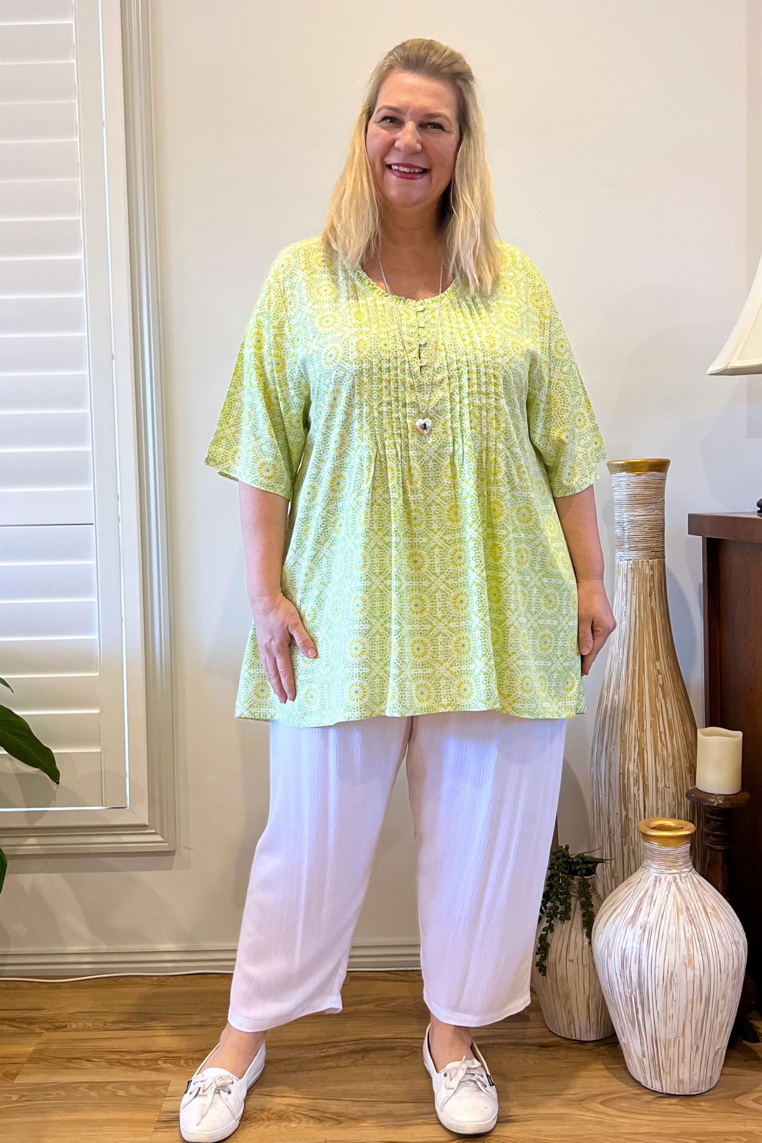 Kita Ku modelling a lime and green pleated Elly top over white pants in a lounge  setting. 
