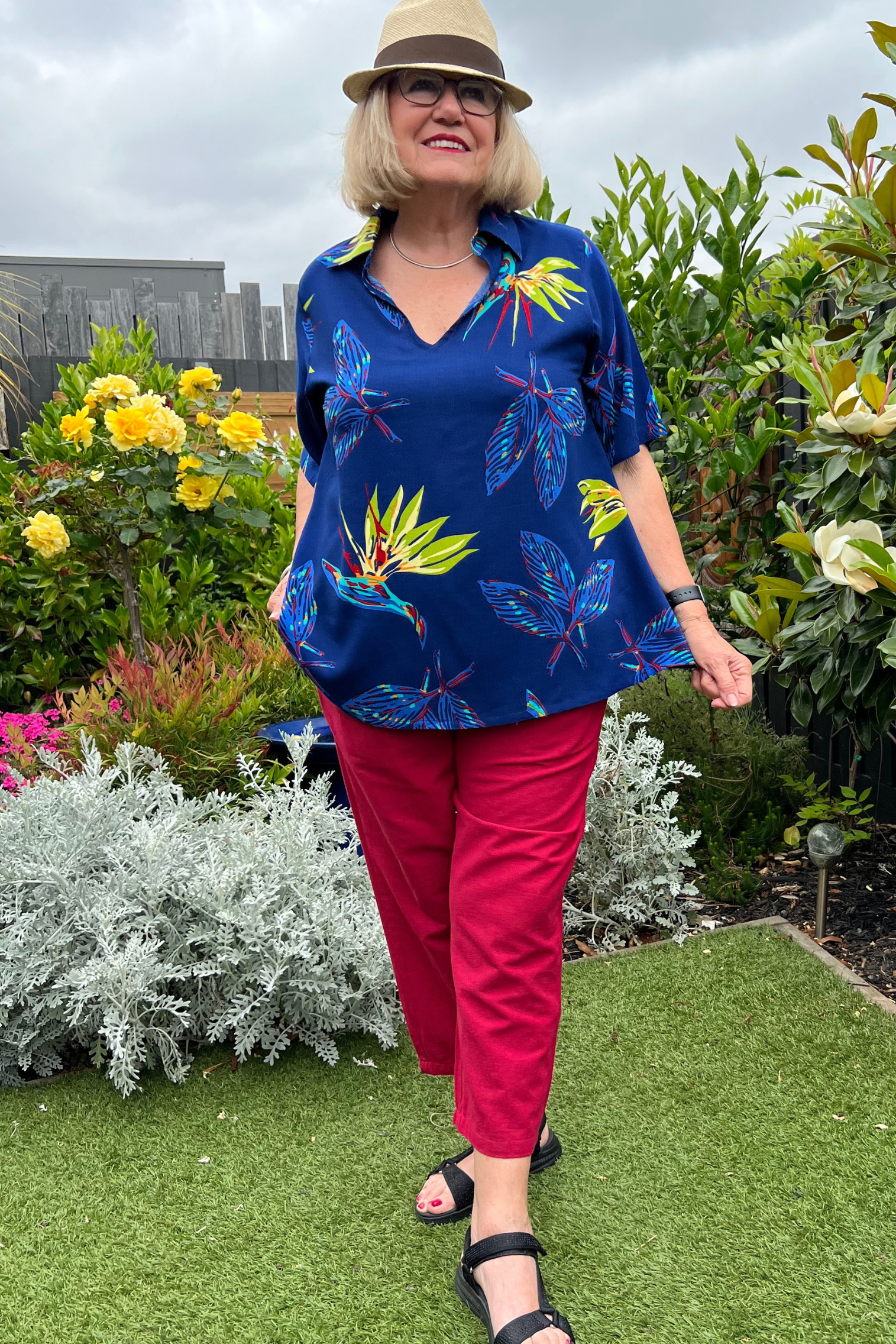 Kita Ku modelling a tropical paradise printed top  Rebecca over red pant port in a garden setting. 