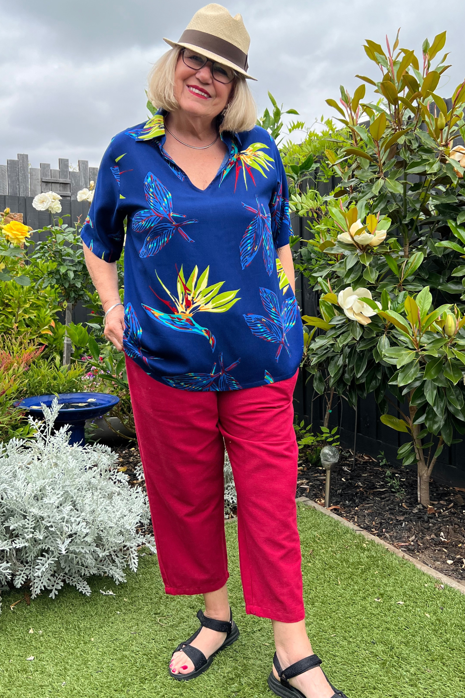 Kita Ku modelling a tropical paradise printed top Rebecca over red pant port in a garden setting.