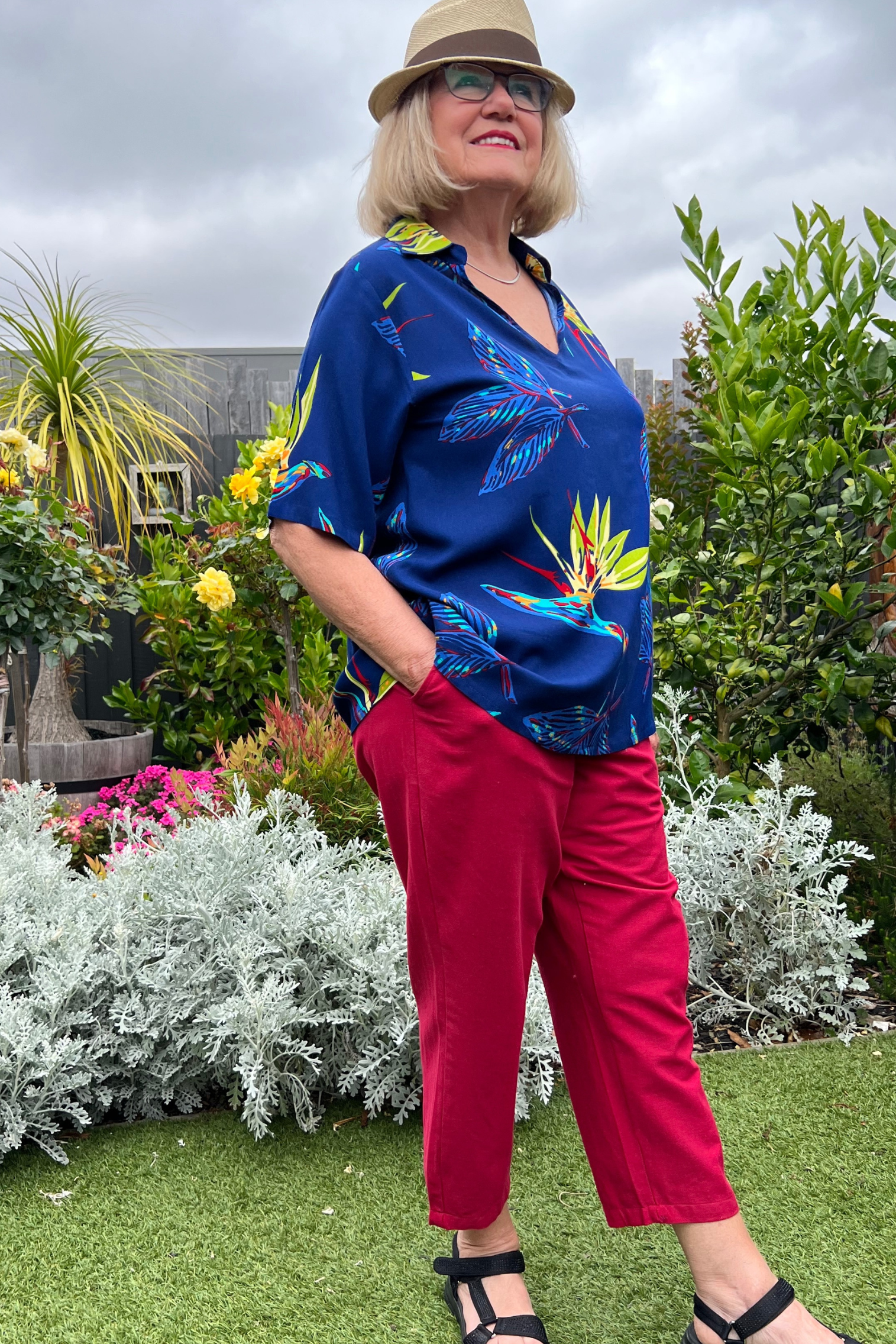 Kita Ku modelling a tropical paradise printed top Rebecca over red pant port in a garden setting.