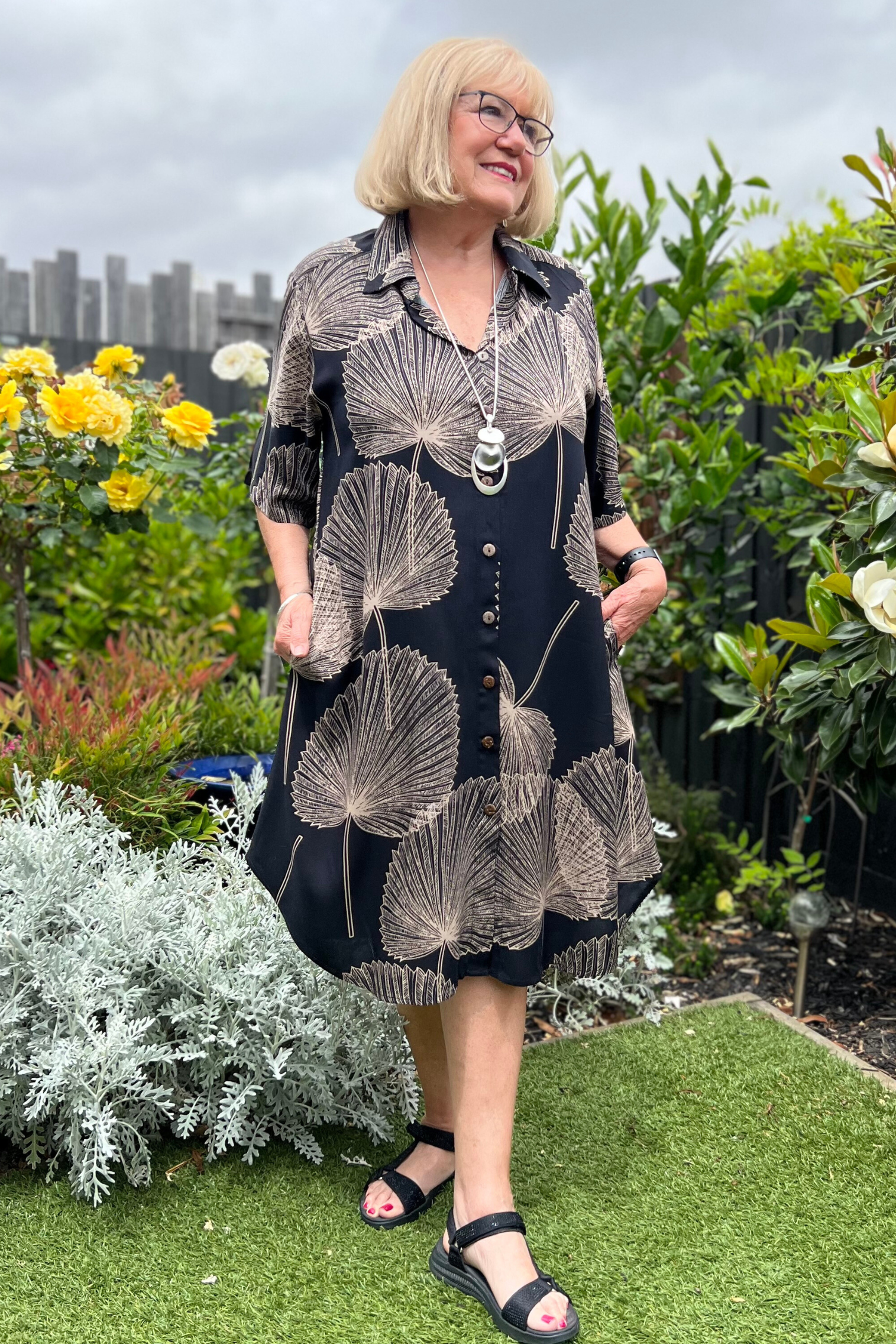 kita ku model in the shirt style dress pronto in a paolo print of fan black and mocha palm leaf set in a sunny garden. 