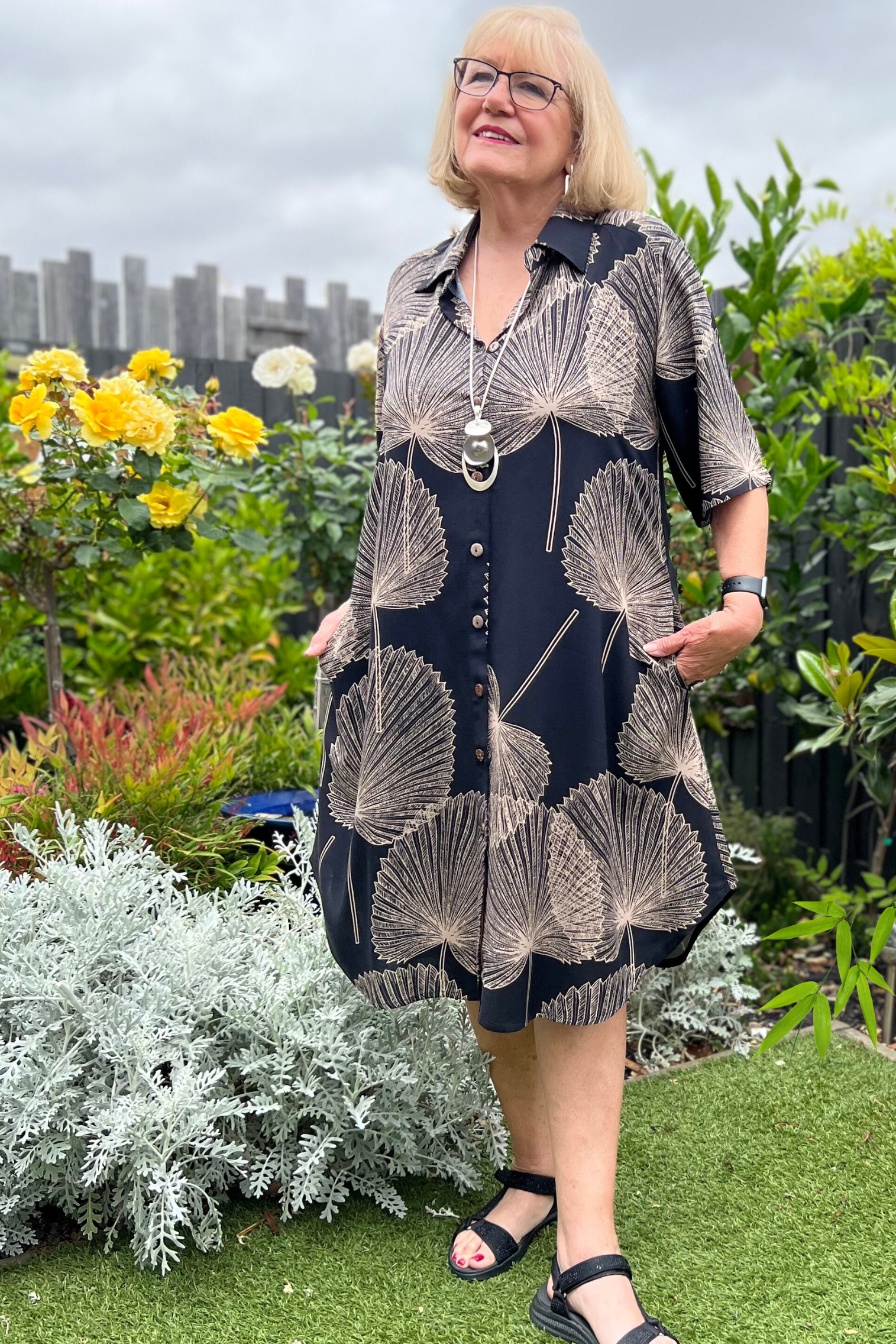 kita ku model in the shirt style dress pronto in a paolo print of fan black and mocha palm leaf set in a sunny garden.