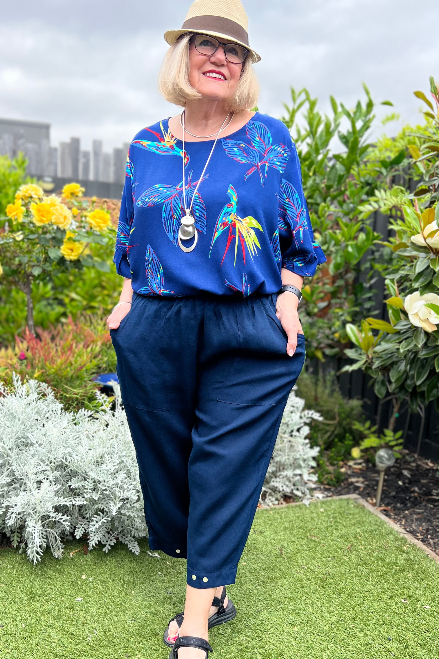 Kita Ku modelling a pant Sita in navy with button trim on the hem, also with a top Zara set in a sunny garden. 