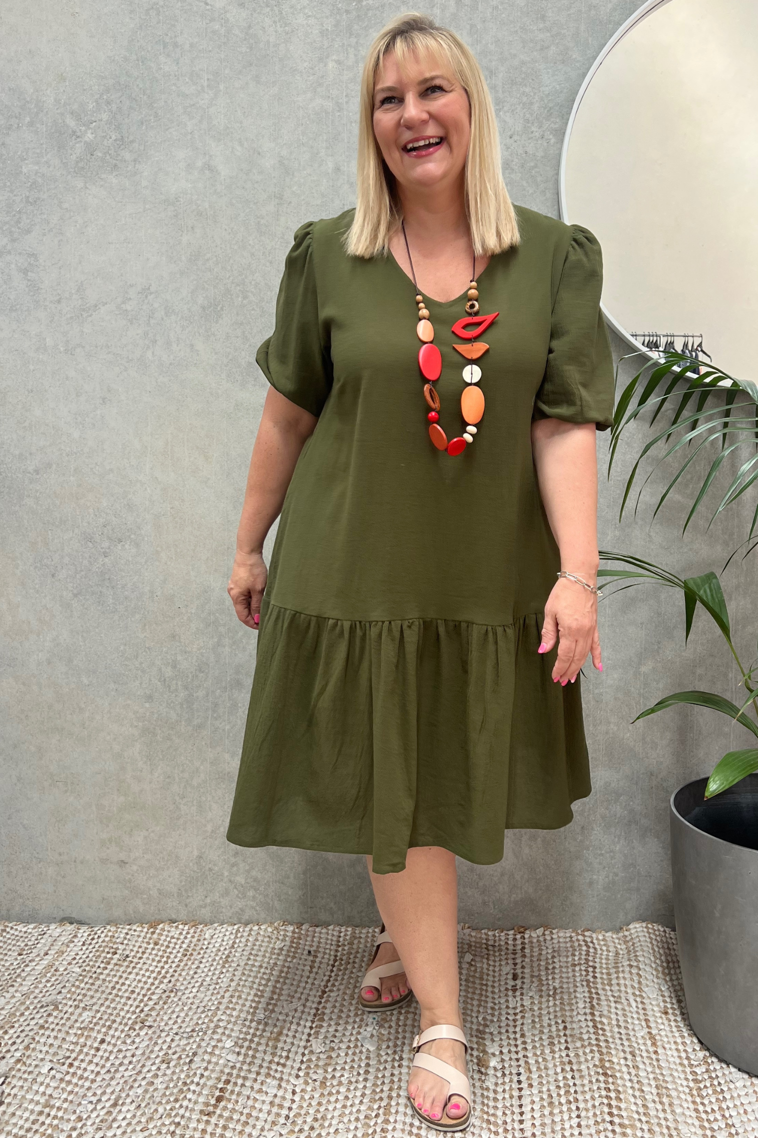 Kita Ku modelling a khaki cotton layer dress and wearing a red necklace in a retail  setting. 