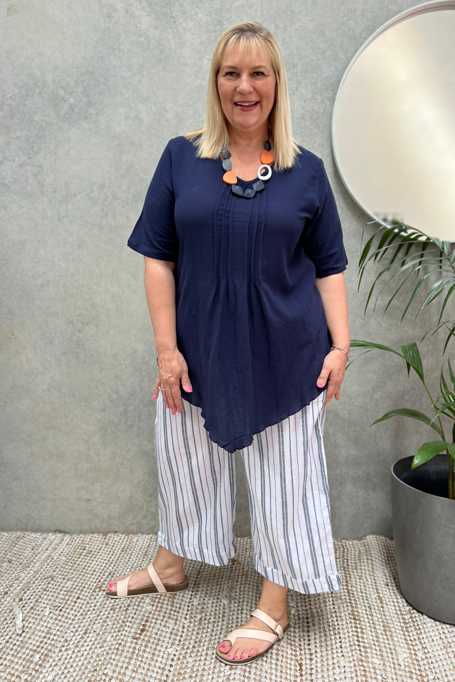 Kita Ku blonde model wearing a navy vee top with pleated front, over a pair of white and navy stripe pants in a retail setting.