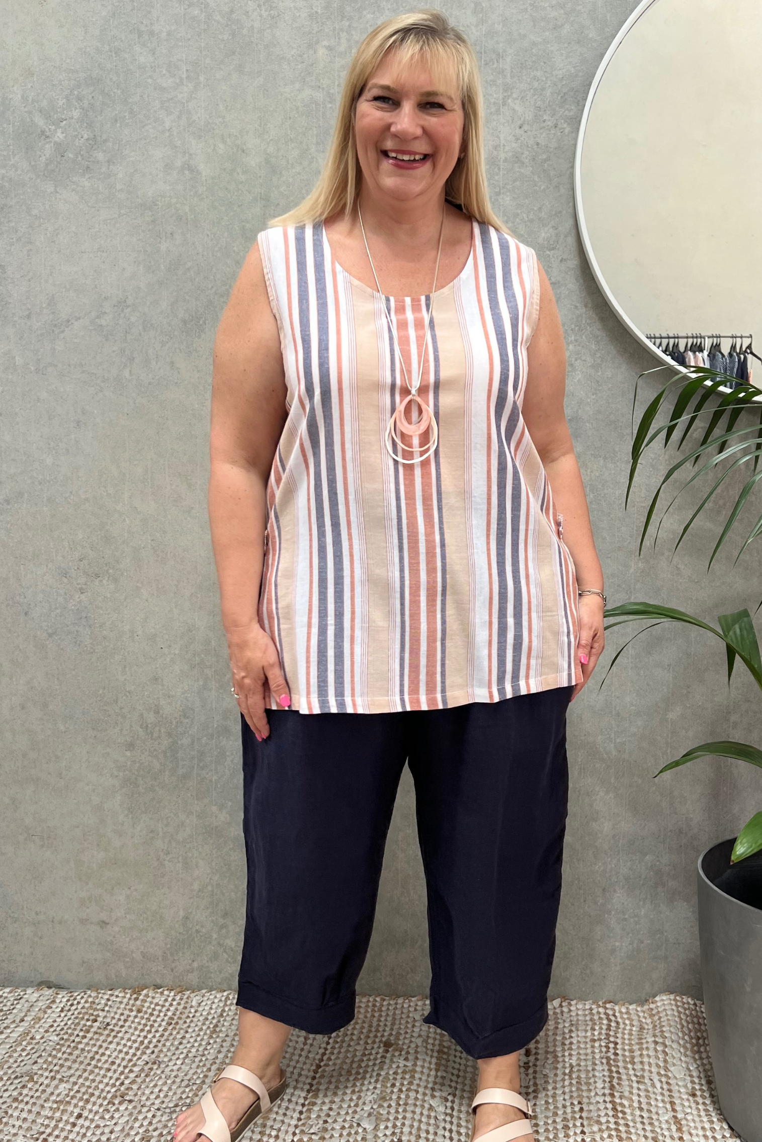 Kita Ku modelling a no sleeve cami of coral and grey stripes over a pant in navy in a retail setting.