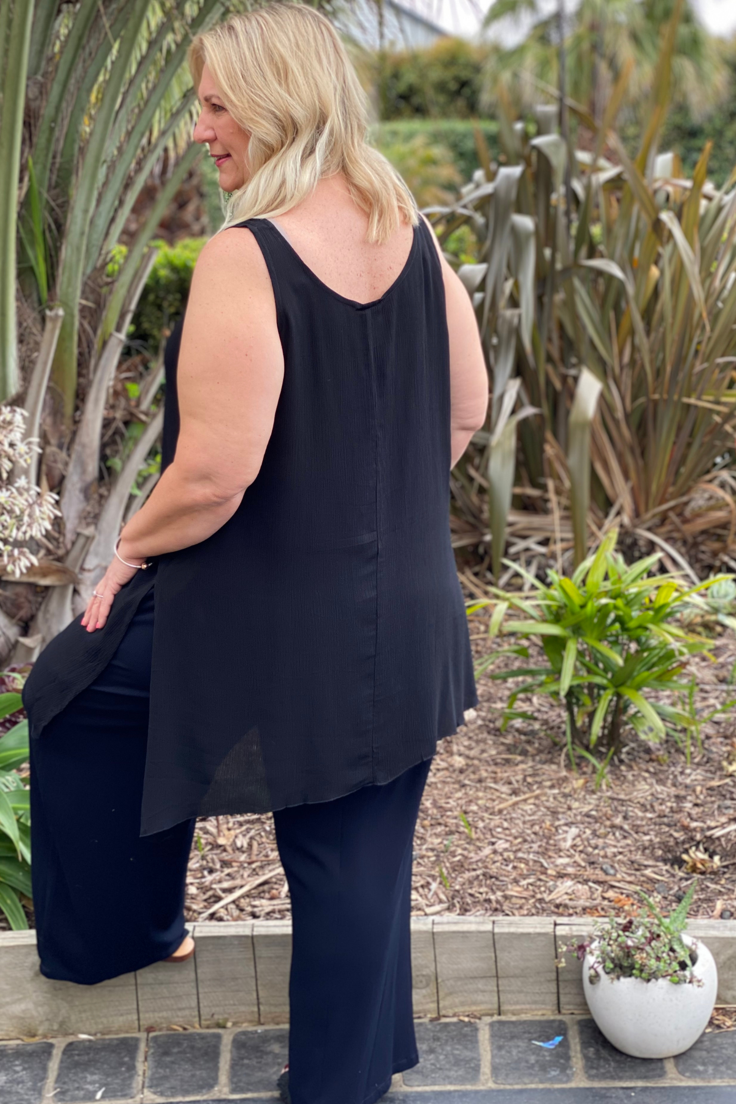Kita Ku model wearing a black asymmetrical longline cami top over black pants in a garden setting.